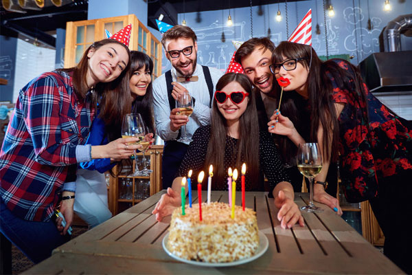 Nhóm bạn đội mũ tiệc vây quanh cô gái đeo kính trái tim ngồi trước bánh sinh nhật cắm nến