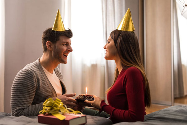 Vợ chồng đội mũ sinh nhật, chồng cầm bánh nhỏ cắm nến, trước mặt là hộp quà thắt nơ