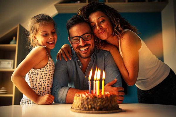 Vợ và con gái ôm người chồng đeo kính đang cười bên bánh sinh nhật cắm nến