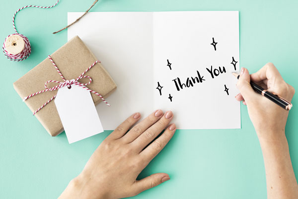 Bàn tay viết chữ Thank You lên tấm thiệp trắng, bên cạnh hộp quà gói giấy kraft buộc dây