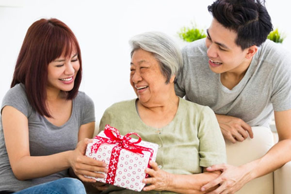 Đôi bạn trẻ tặng hộp quà thắt nơ đỏ cho người mẹ tóc bạc đang cười rạng rỡ
